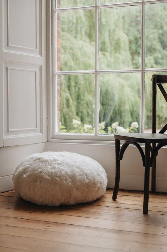 White sheepskin footstool on a wooden floor with a large window in the background showing greenery.