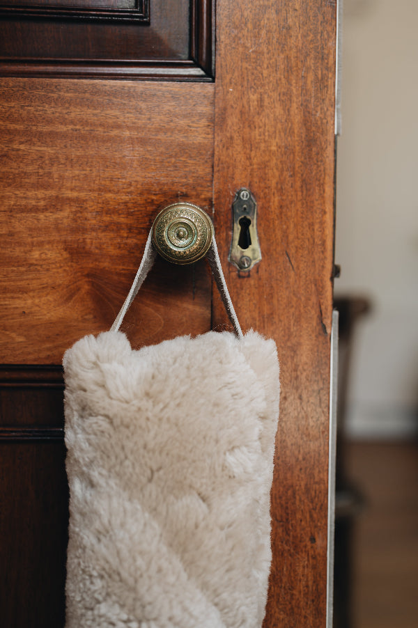 Oatmeal sheepskin draught excluder with leather handle hanging on a wooden door with a brass doorknob and keyhole.