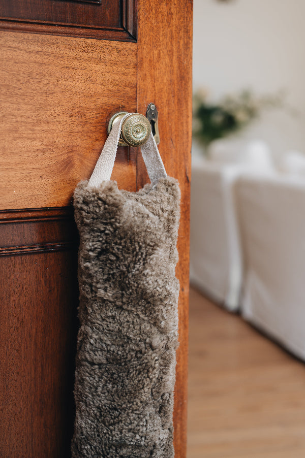 Wheat brown sheepskin draught excluder with leather handle hanging on a wooden door with a brass doorknob and keyhole.