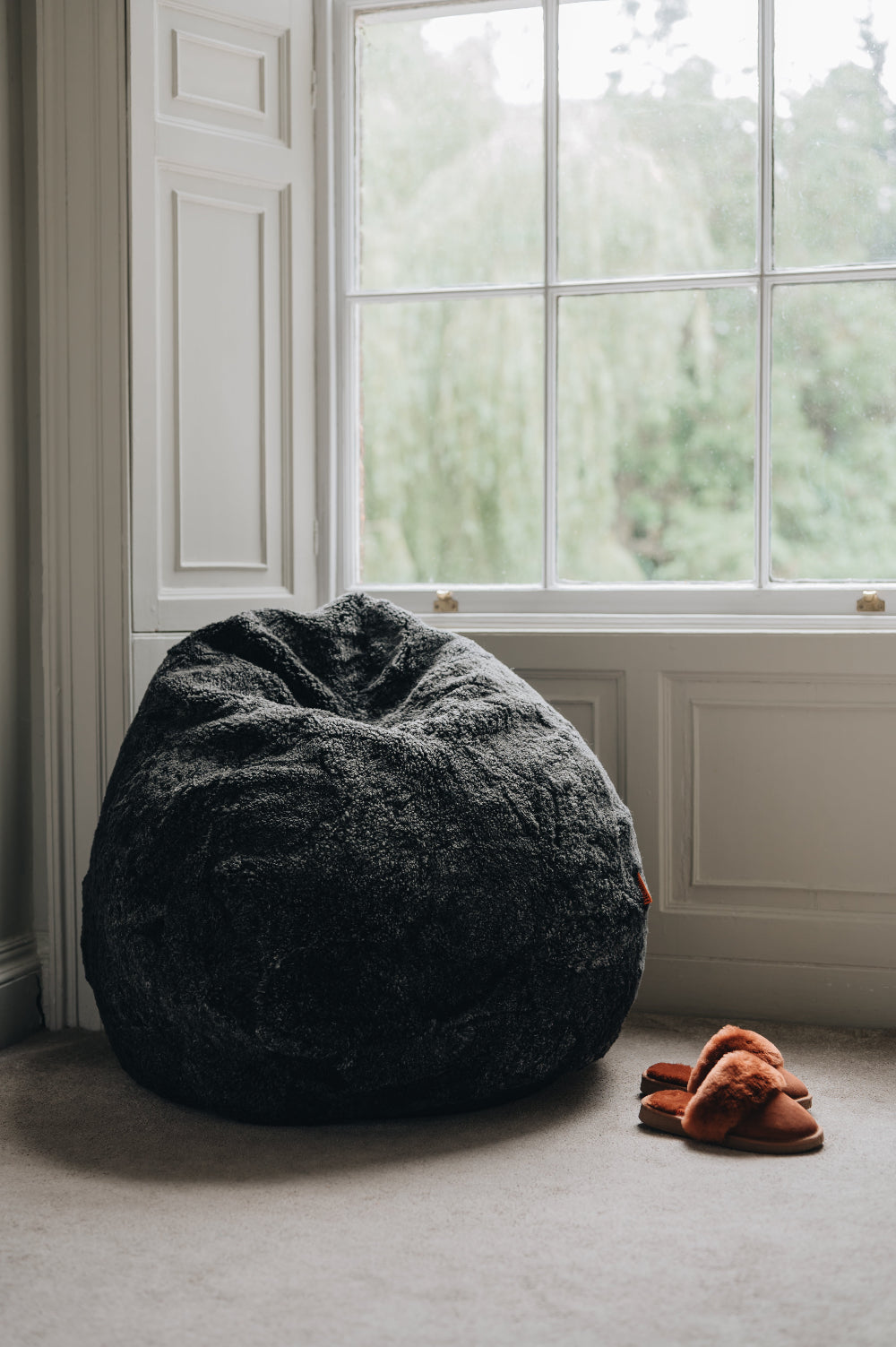 Black Smoke sheepskin bean bag chair next to a window with a pair of slippers on a carpeted floor.