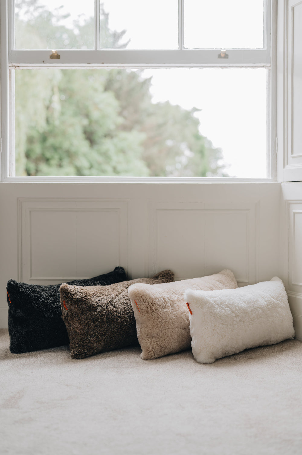 Four sheepskin cushions in black, brown, beige, and white on a light-coloured carpet with a window in the background.