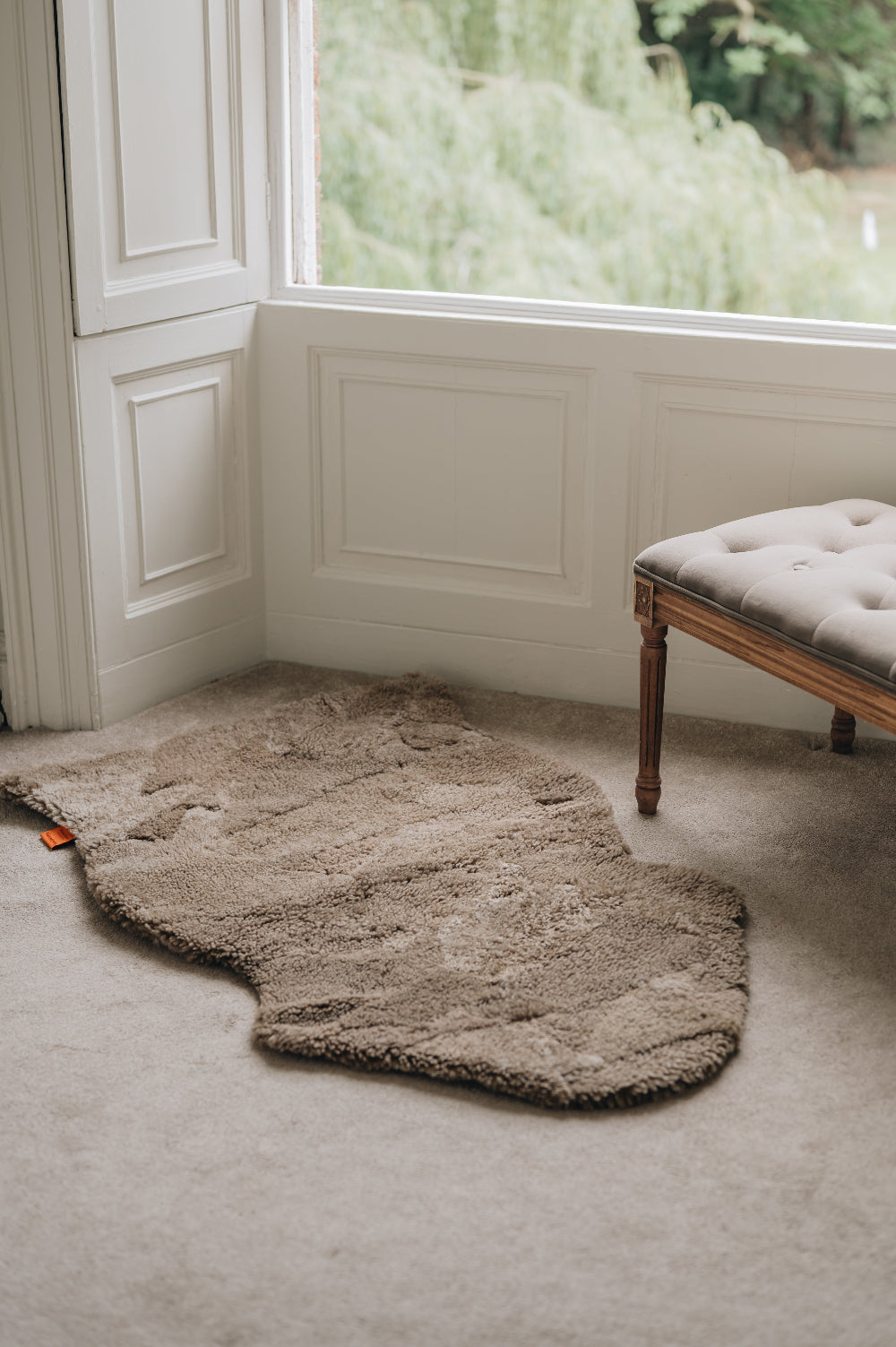 Wheat brown sheepskin rug on a carpeted floor with a window and bench in the background