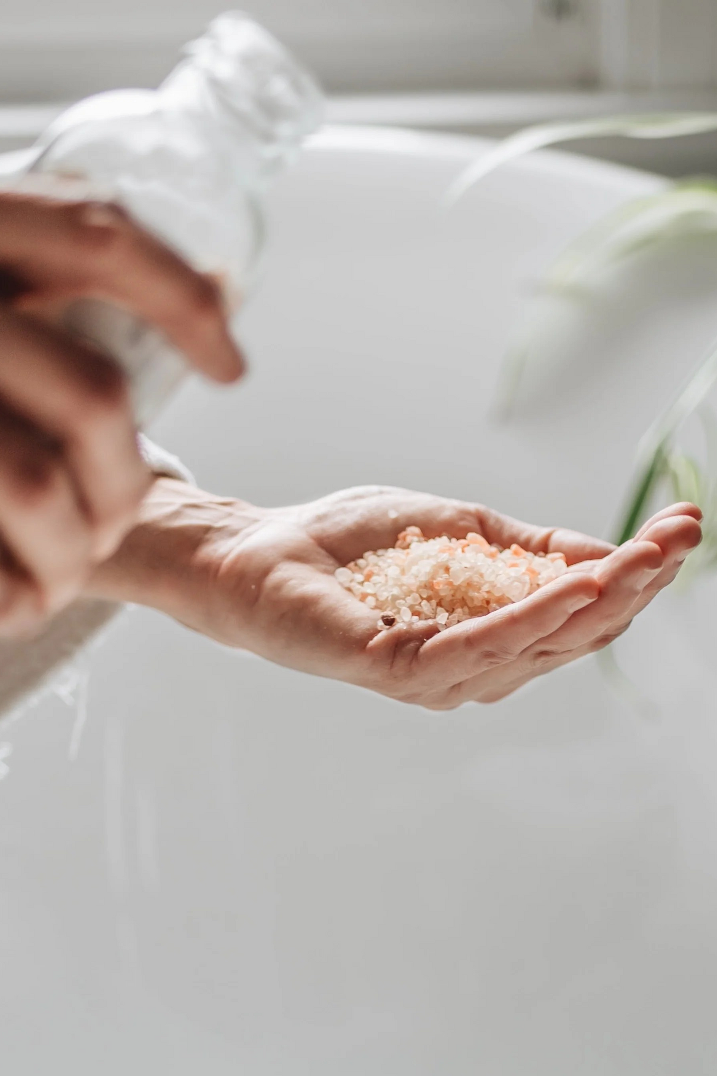 Pink and white bath salts being held in someones hand over a white bath, the glass bottle container for them being held in the foreground