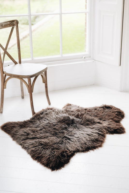 Soft Natural Brown British Sheepskin Rug contrasting against a hard and stark white wooden floor next to a traditional wooden chair in a window.