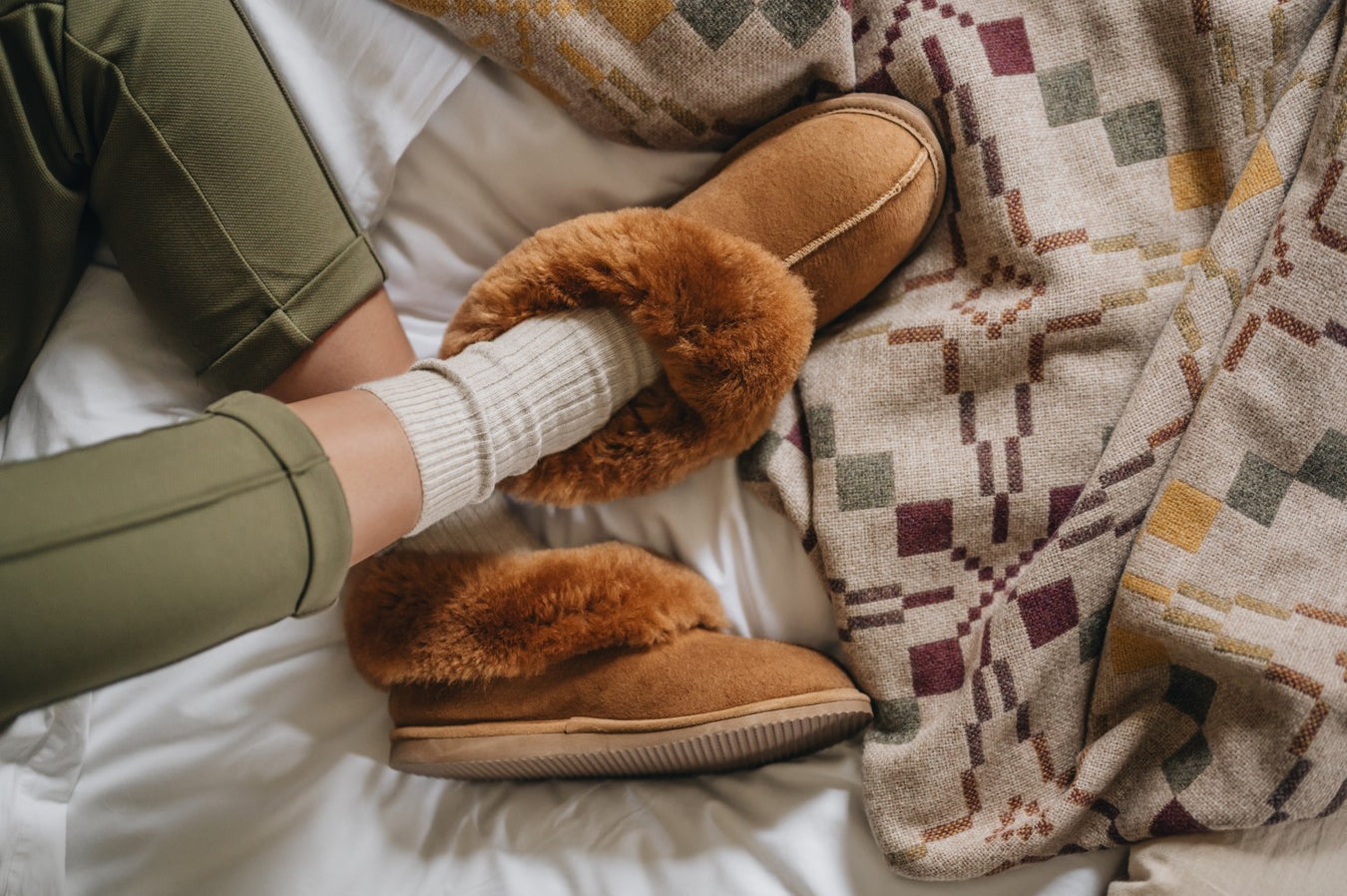 top view of warm brown/orange sheepskin slipper boots on a bed. Featuring a fluffy trim, soft suede outer, and decorative centre seam