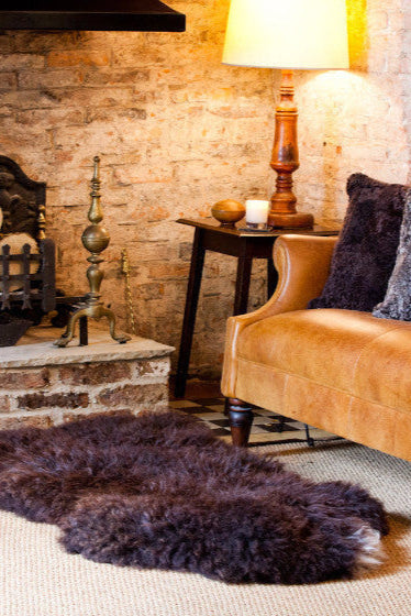 Cosy Brown British Sheepskin Rug, in front of the fire on the floor with accent Sheepskin cushions on a leather sofa next to it.