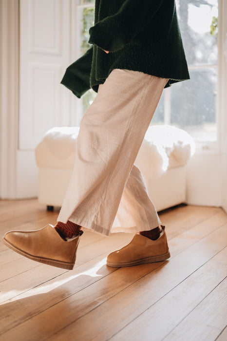 Young woman wearing  tan sheepskin slipper boots and beige trousers indoors on a wooden floor.
