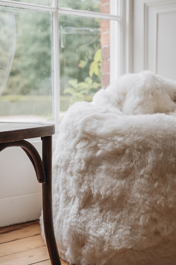 Close up of a white sheepskin bean bag next to a wooden chair next to a window with a view of greenery