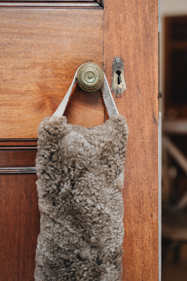 Wheat brown sheepskin draught excluder with leather handle hanging on a wooden door with a brass doorknob and keyhole.