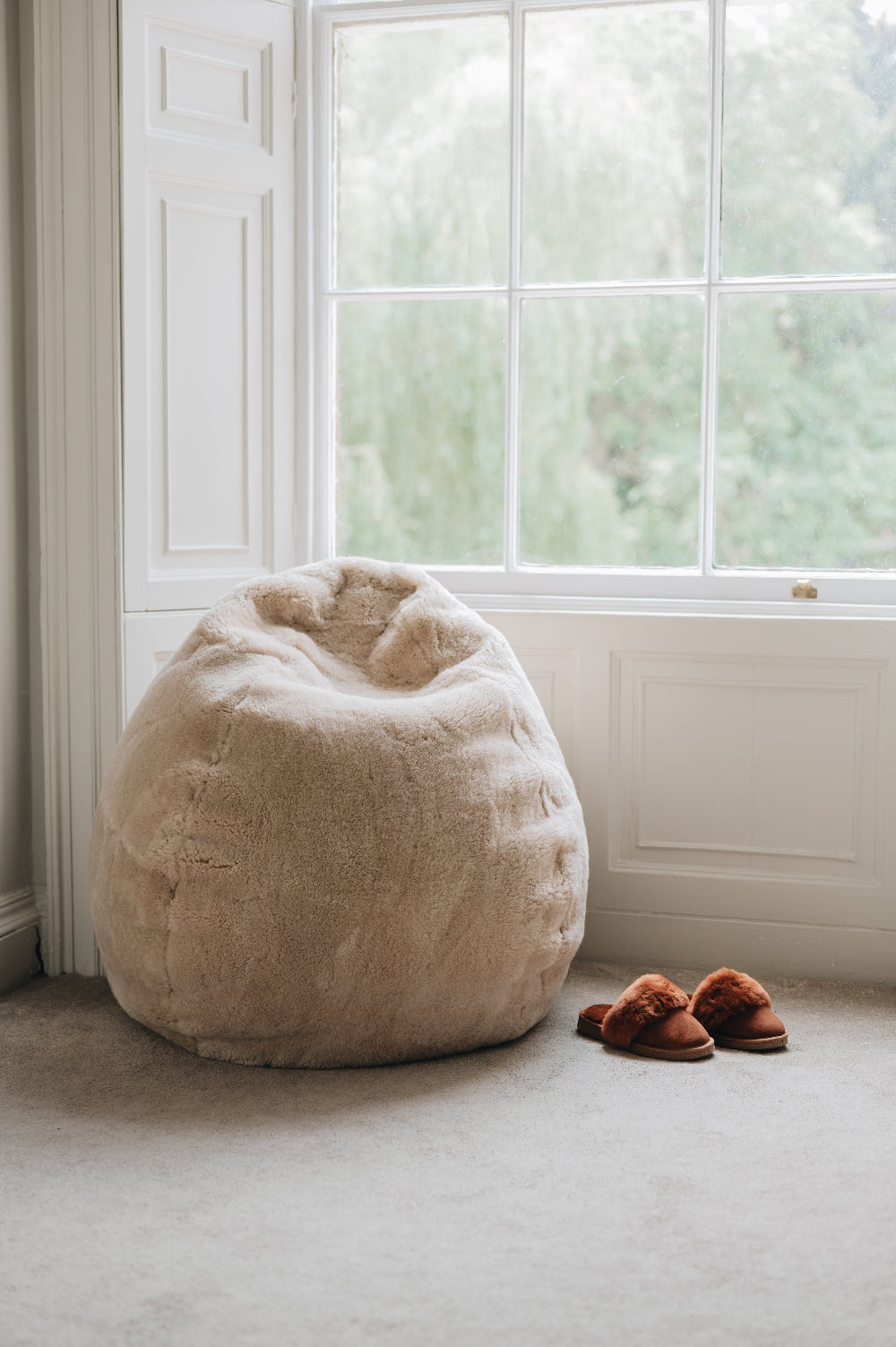 Oatmeal sheepskin bean bag chair next to a window with a pair of slippers on a carpeted floor.