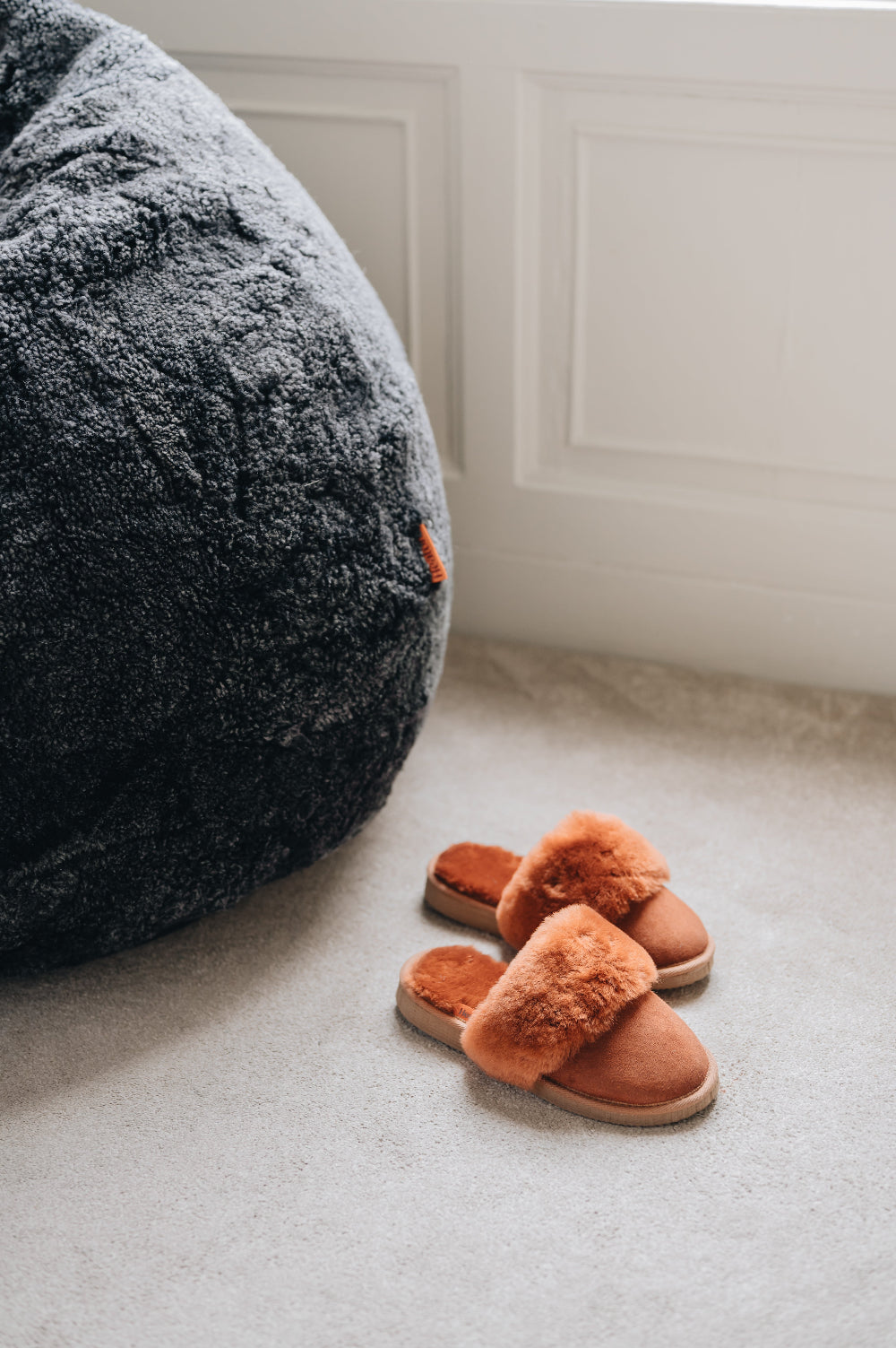 Pair of rust coloured sheepskin slippers on a light  carpet next to a large dark sheepskin bean bag chair.