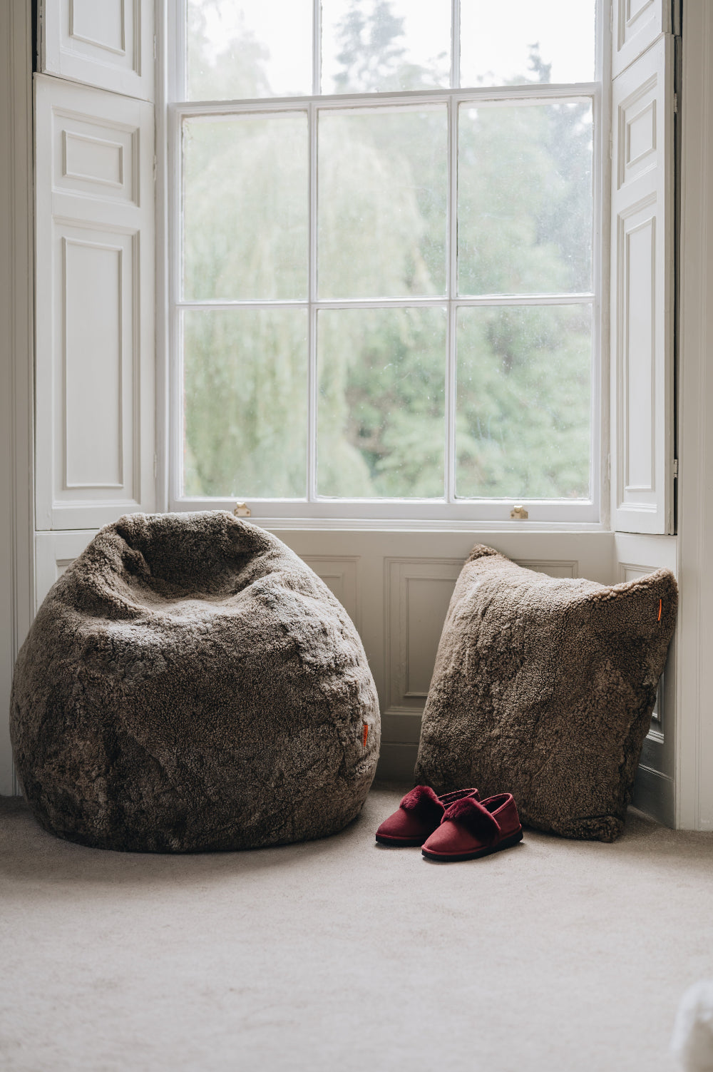A wheat brown sheepskin beanbag and floor cushion on a light carpeted floor in front of a large window with a view of greenery.