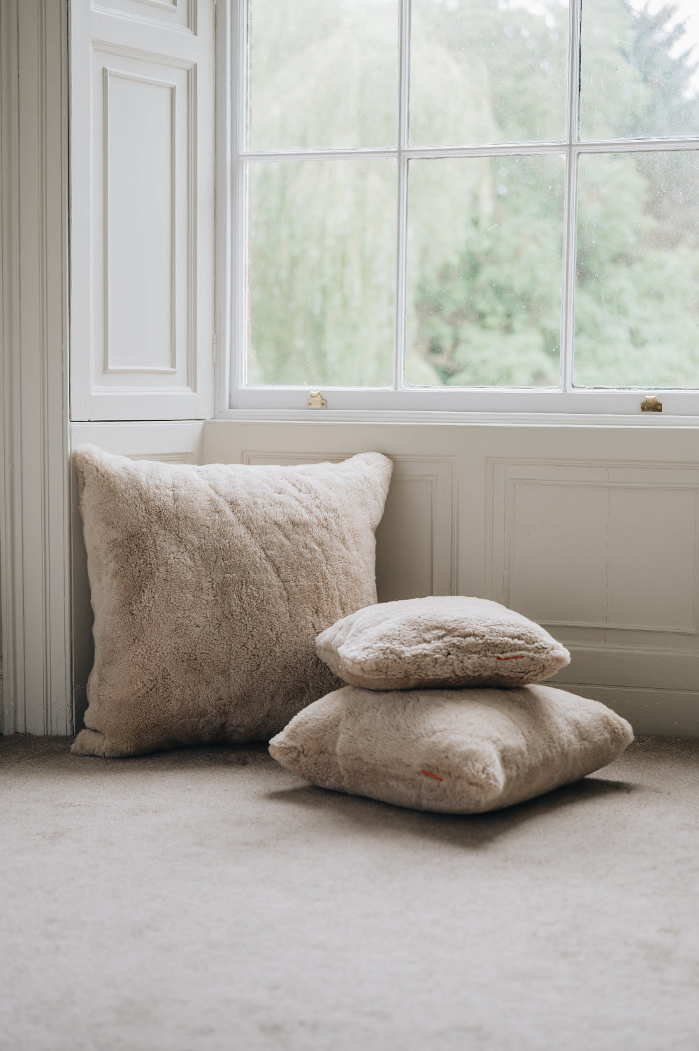 Oatmeal floor cushion plus 50cm and 40cm cushions on on a carpeted floor in front of a window with a white frame.