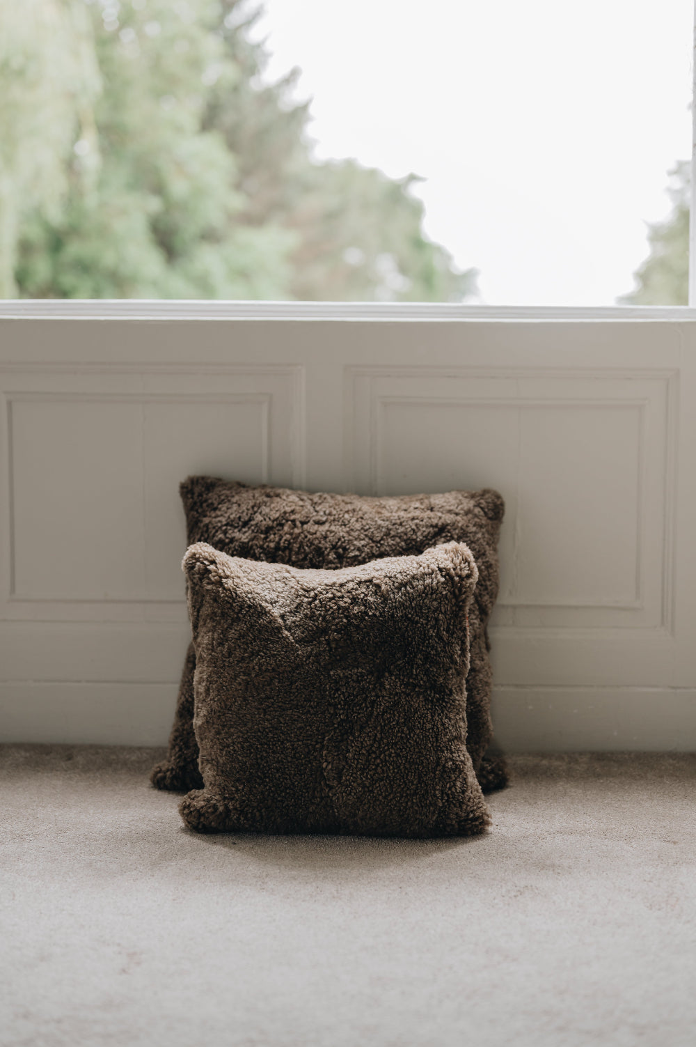 Two wheat brown sheepskin cushions on a light-coloured floor with a blurred window and greenery in the background.