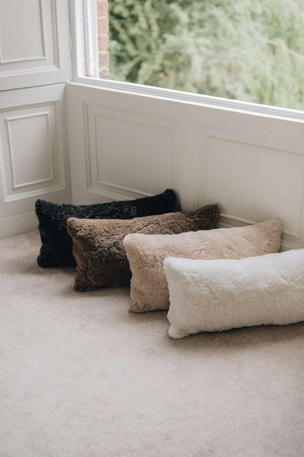 Four rectangular sheepskin cushions in black, brown, beige, and white on a carpeted floor near a window.