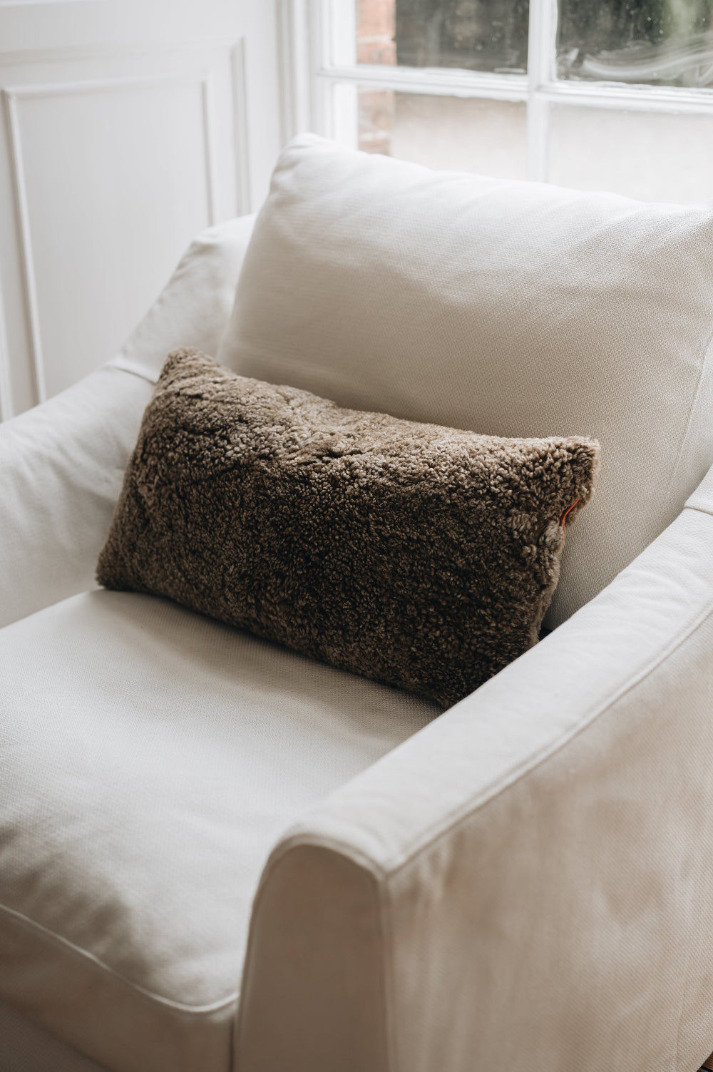White armchair with a brown textured sheepskin cushion in a softly lit room.