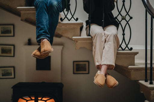 Man and women sitting with legs hanging over the edge of a staircase. The man is wearing the Henry slipper and the woman is wearing the Tilly Slipper, both with their ankles crossed.