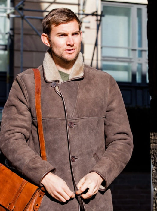 Model in Edward Brown Sheepskin Men's Coat, wearing a brown leather satchel, walking down hill.