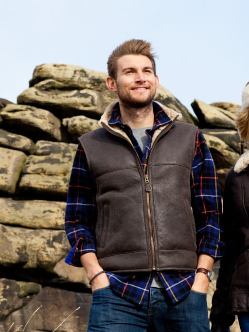 Men's Sheepskin Gilet, with collar, frotn zip and pockets. Male model stands near rocky backdrop.