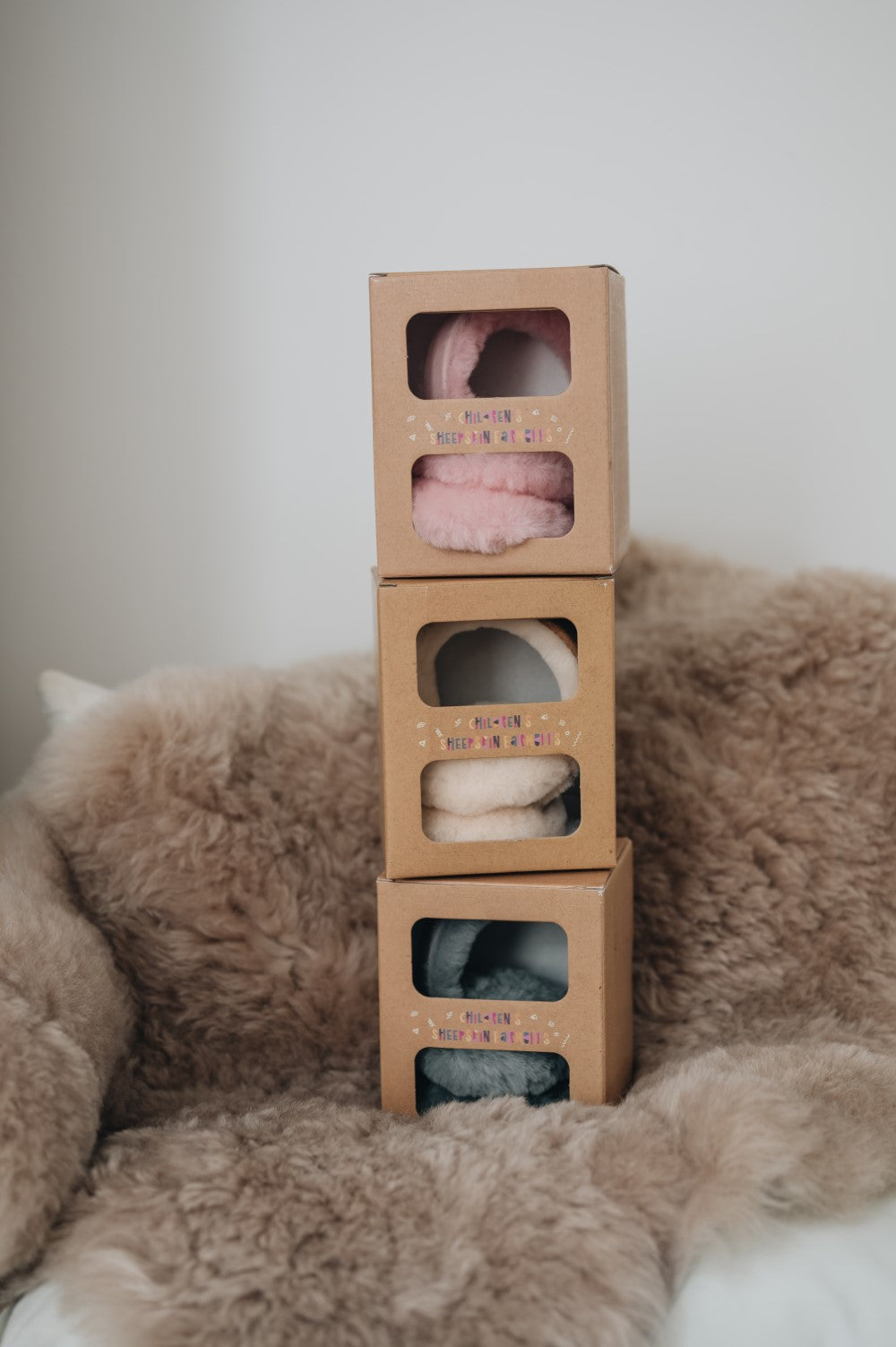 three stacked boxes of children's sheepskin earmuffs in pink, grey and tan
