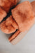 Close-up of a pair of fluffy rust coloured sheepskin slippers with an elasticated band and an orange cork Heatons brand label on a light background