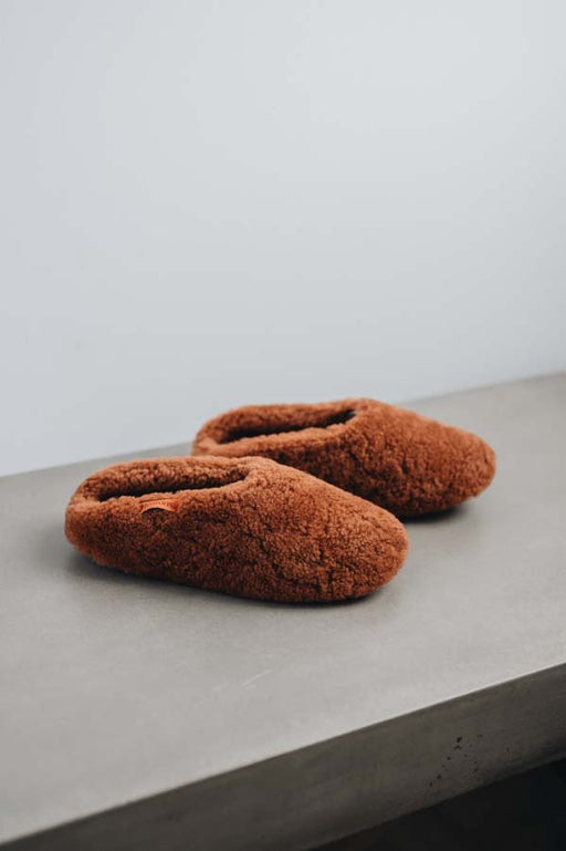Pair of rust coloured curly sheepskin slippers on a concrete surface with a light grey background