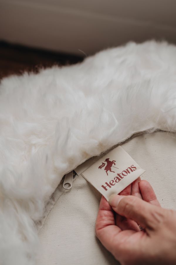 Hand holding a 'Heatons' tag on the bottom of a white sheepskin footstool