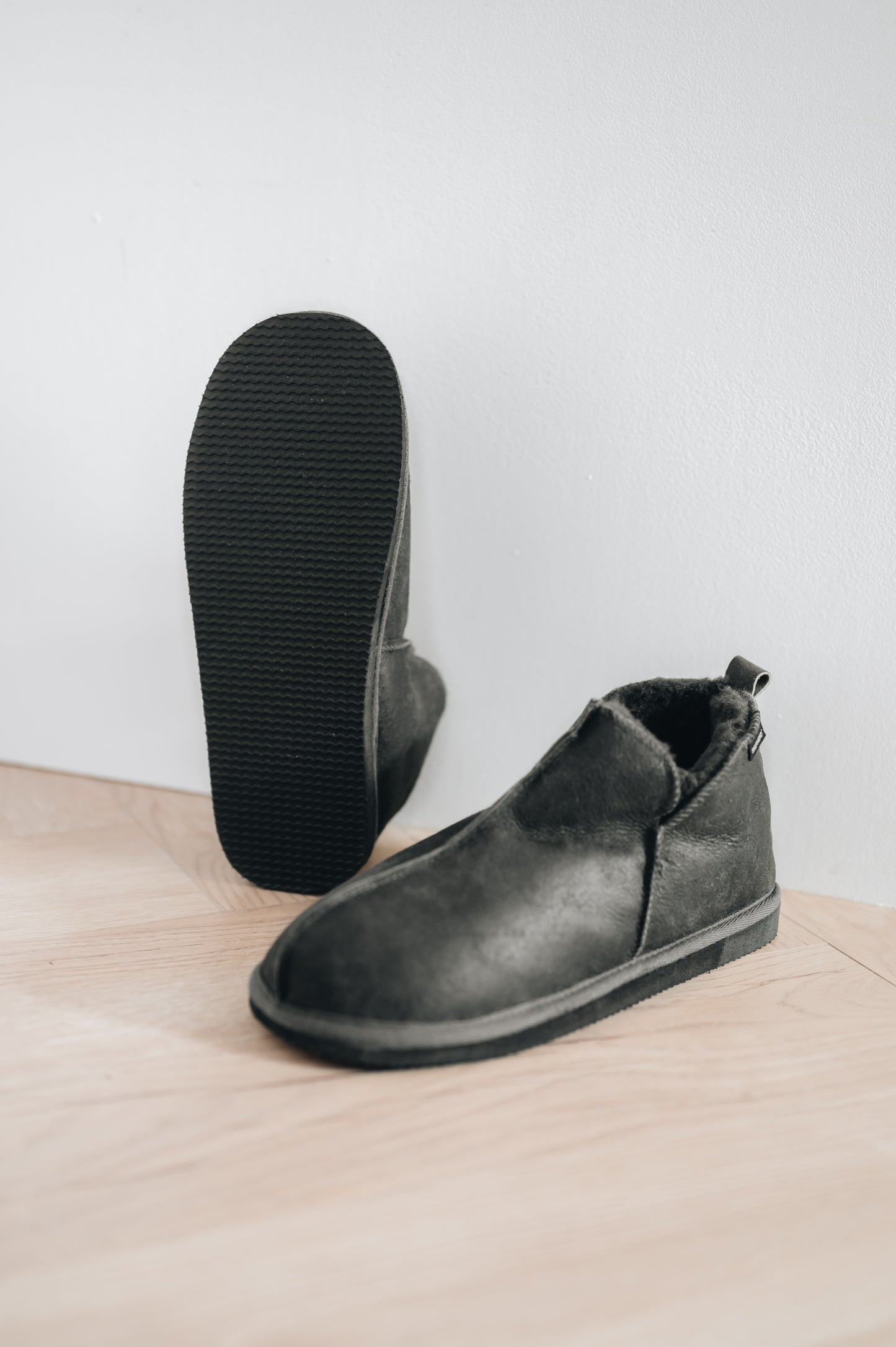 men's sheepskin slippers in dark grey, right slipper propped up against wall to show off EVA foam sole
