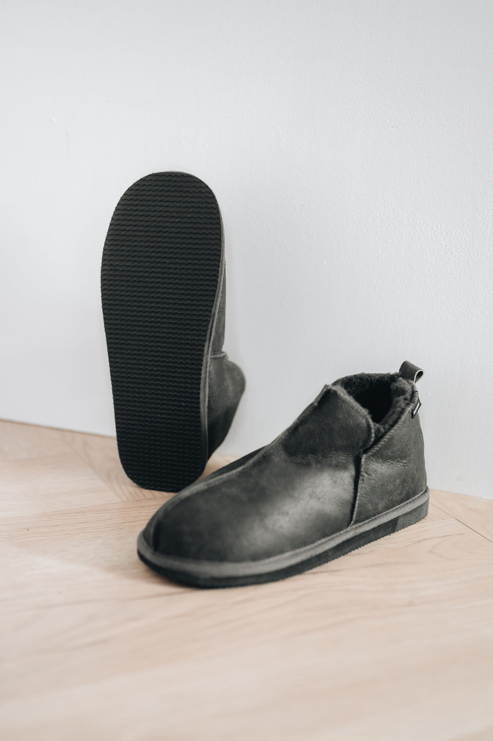 men's sheepskin slippers in dark grey, right slipper propped up against wall to show off EVA foam sole
