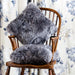 Icelandic Sheepskin Cushion in Grey, on wooden chair against floral background.