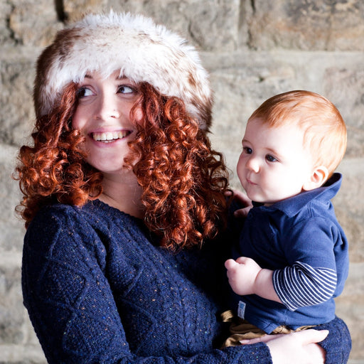 An auburn curly hair model wears a navy blue cable knit jumper. She is smiling and holding a child. She wears a White and Brown Natural Full Sheepskin headband. The headband covers her ears and forehead.
