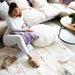 Wide view of Icelandic White Patchwork Sheepskin Melange Rug in a living room. Features white, beige and grey coloured patches. A woman sits on top of the rug with Gabriella beige slippers and holding a Melin Tregwynt purple patterned cushion.