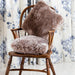 Icelandic Sheepskin Cushion in Taupe, on wooden chair against floral background.